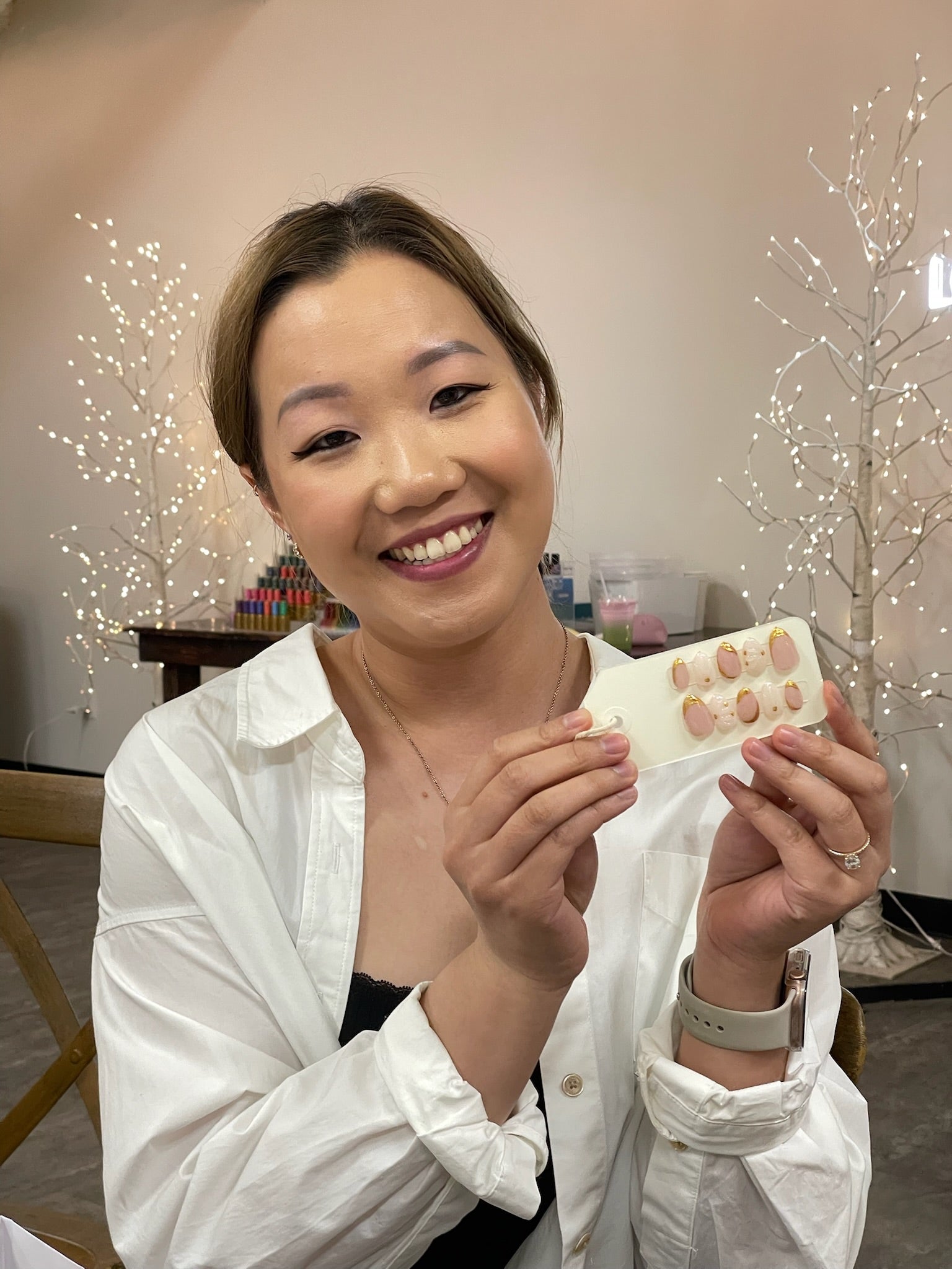 Woman holding a set of painted press-ons, smiling in a room with decorative lights.