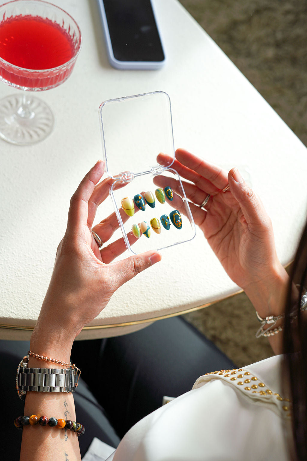 Person holding a press-on nail case with colorful designs on a table with a drink and phone.